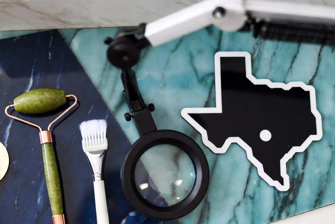Flat lay of esthetician tools including jade roller, facial brush, magnifying lamp, and a Texas-shaped graphic element